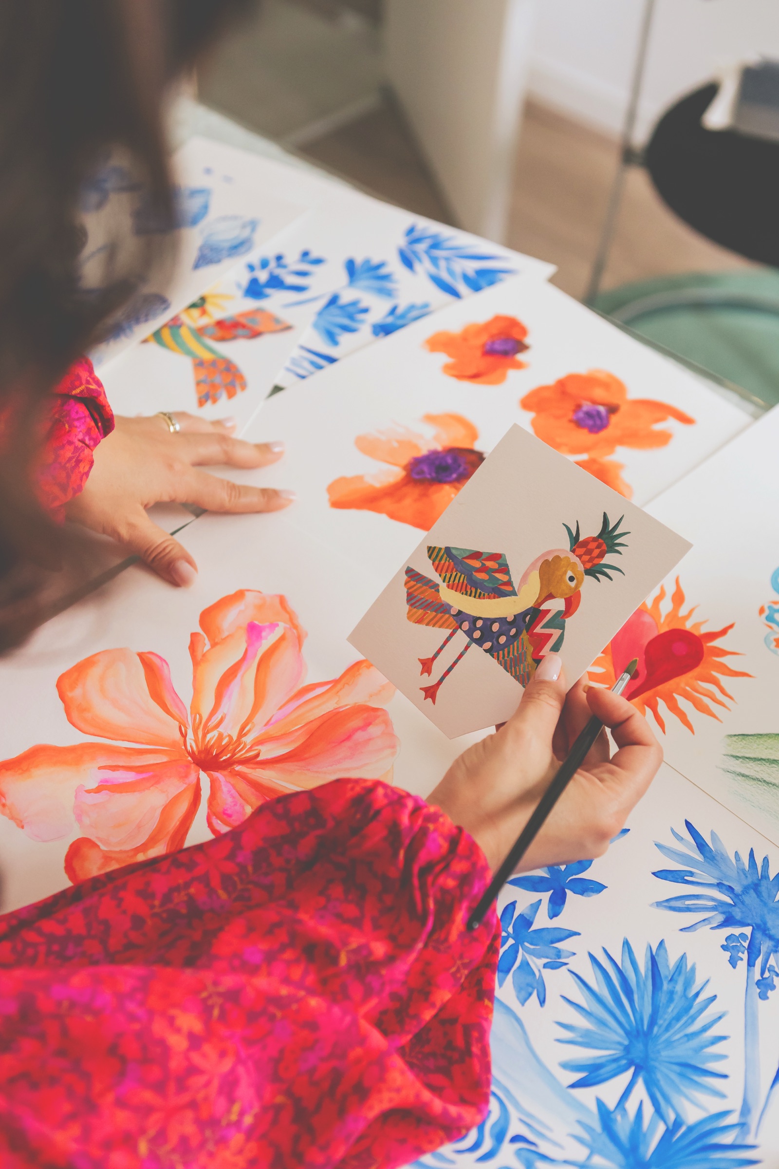 Anna holding a painted folk-art bird over painted florals spread across the studio table