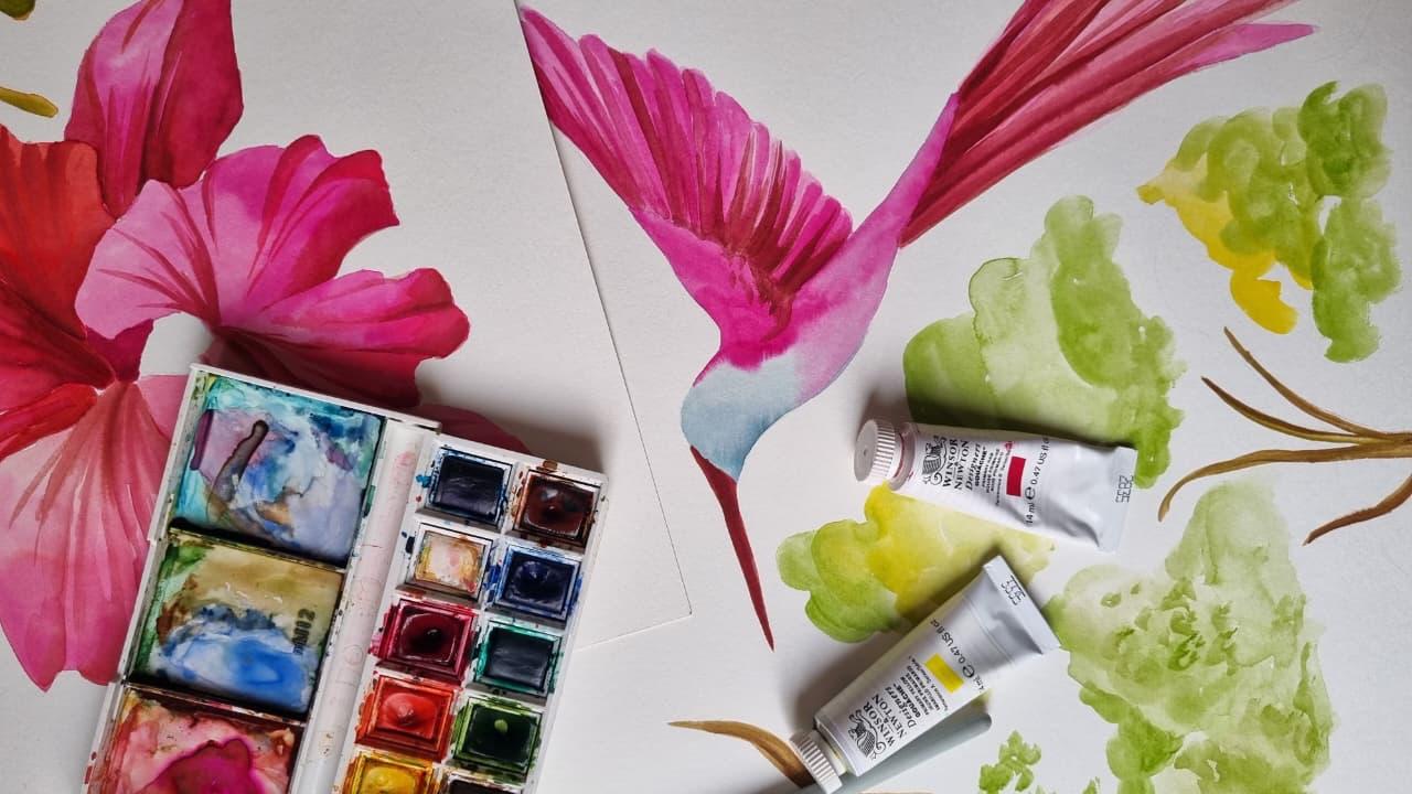 A hummingbird and hibiscus painted in watercolor beside the open pan set and paint tubes